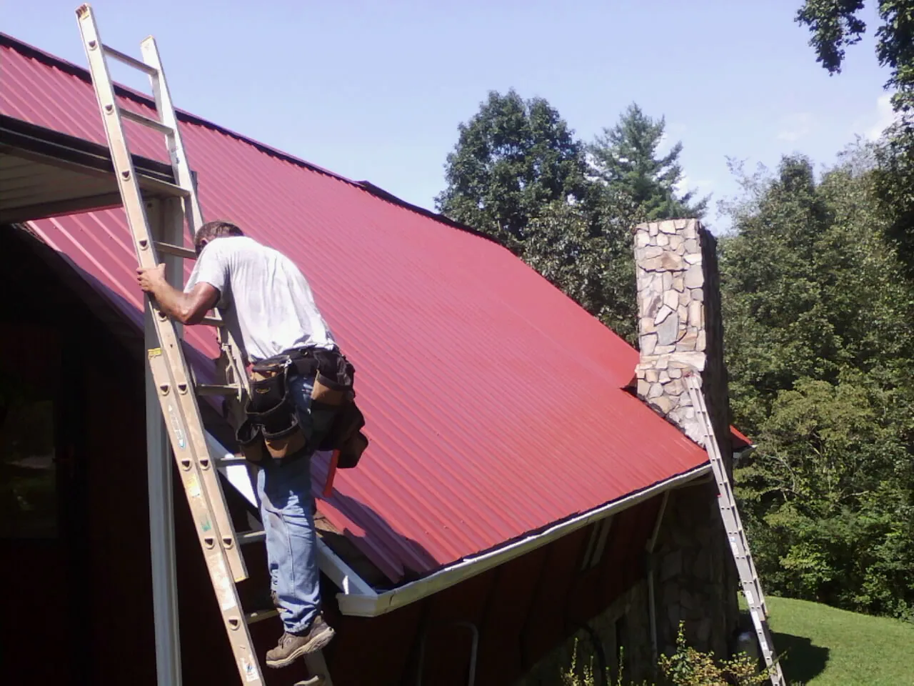 Roofer on a ladder beside a red metal roof and stone chimney — Soffit & Fascia Repair by Roofing Brothers in North Wales